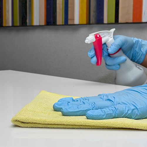 A person wearing blue gloves is cleaning a surface with a yellow cloth and a spray bottle in hand.