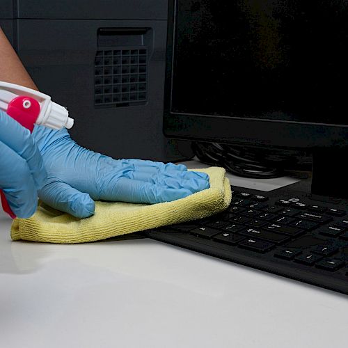 A gloved hand is spraying and wiping a keyboard and desk area with a yellow cloth, indicating a cleaning process near a computer.