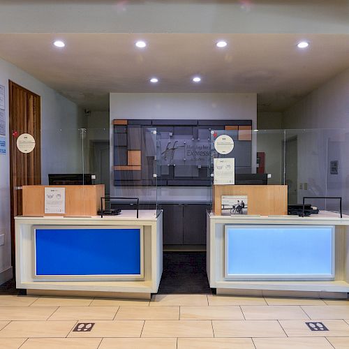 The image shows a reception area with two desks, a glass partition, blue accents, and a hand sanitizer station to the right.