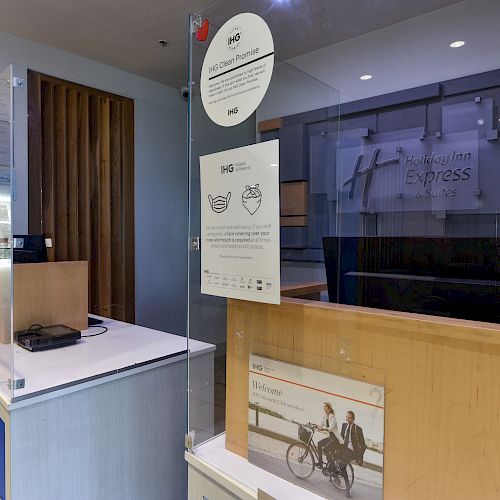 The image shows a hotel reception area with a signboard, a counter, and protective glass. There's a pamphlet holder and signage for 