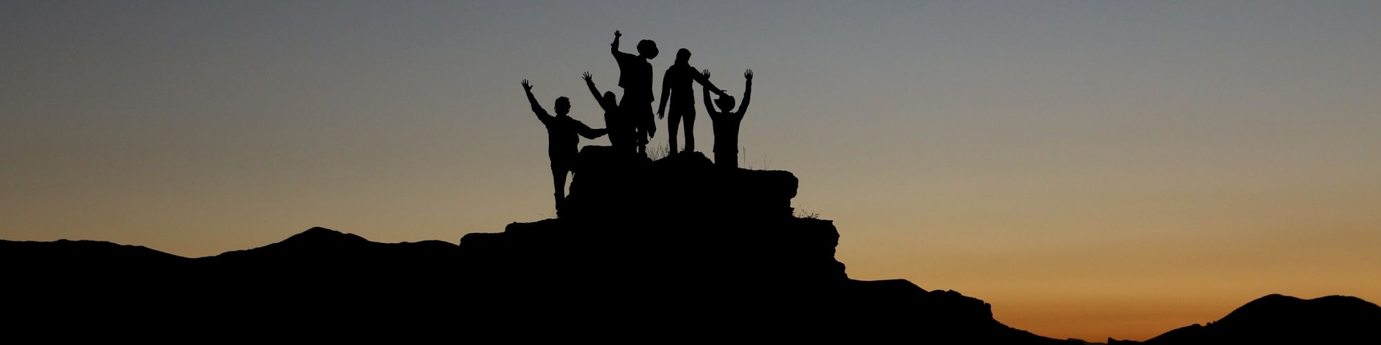 A group of people is silhouetted against a sunset or sunrise, standing on a rocky formation with their hands raised in celebration.