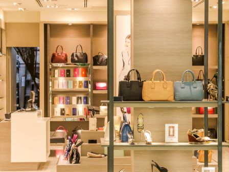 A well-lit boutique displaying handbags, shoes, and accessories. Shelves are neatly organized with various colorful merchandise and a checkout counter.