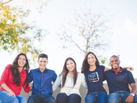 Five people are sitting outdoors on a ledge, smiling and posing for the camera in casual clothing.