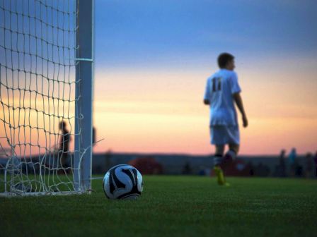 A soccer ball is near a goal post on a field, with players in the background during sunset.