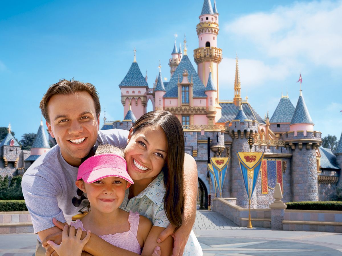 A smiling family of three, with a child wearing a pink cap, poses in front of a castle at what appears to be a theme park on a sunny day.