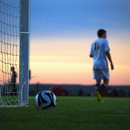 A soccer player stands near the goalpost with a ball on the ground during a game at sunset.