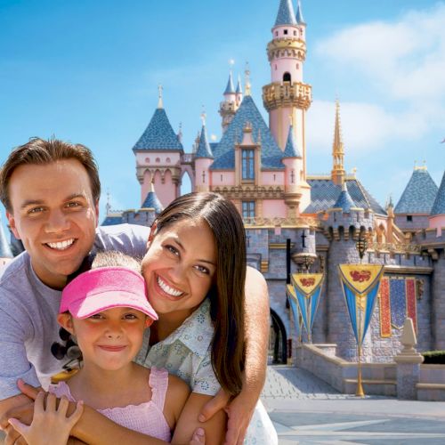 A family of three smiles in front of a castle at a theme park, with blue skies and colorful banners in the background.