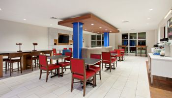 The image shows a modern indoor dining area with red chairs, tables, a TV on the wall, and a counter with a coffee machine and supplies, brightly lit.