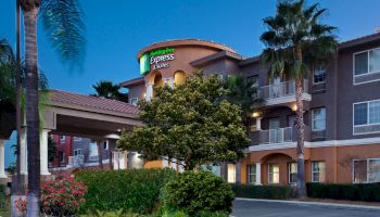 The image shows the exterior of a hotel building, identified as a Holiday Inn Express & Suites, surrounded by palm trees and greenery at dusk.