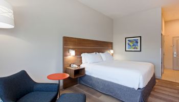 A modern hotel room with a neatly made bed, bedside lamps, an armchair with an ottoman, a small round table, framed artwork, and a lit entryway.