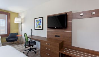 A modern hotel room with a bed, desk with chair, wall-mounted TV, abstract artwork, floor lamp, and an armchair by the window.