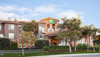 The image shows a hotel building with a landscaped entrance, surrounded by trees and bushes under a clear blue sky.