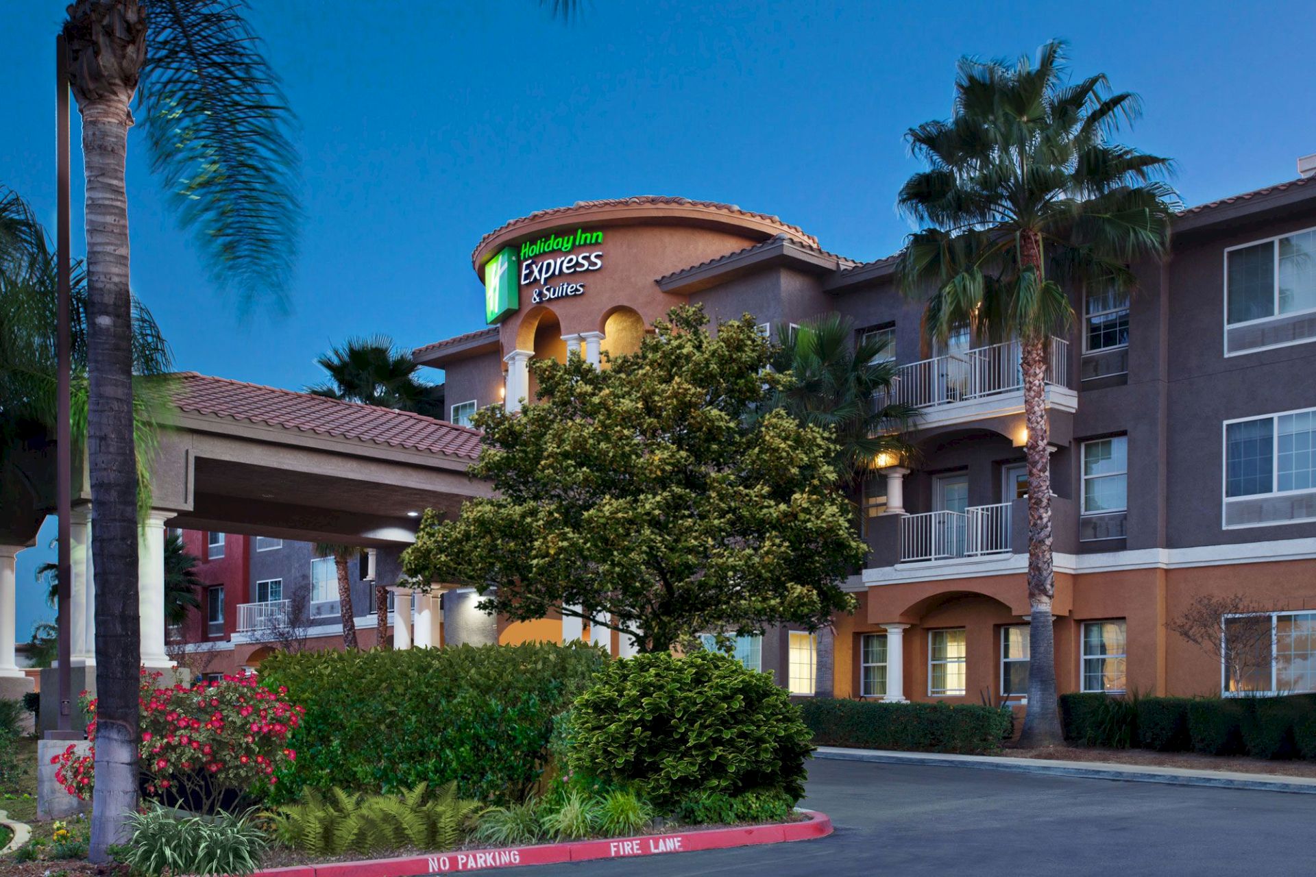 This image shows the exterior of a hotel with palm trees, greenery, and a well-lit entrance during the evening.