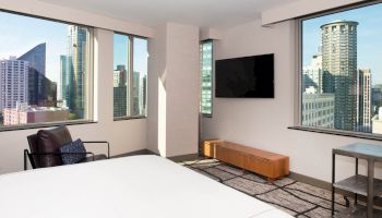 A modern hotel room with a bed, armchair, wall-mounted TV, and large windows showcasing an urban cityscape and skyscrapers.