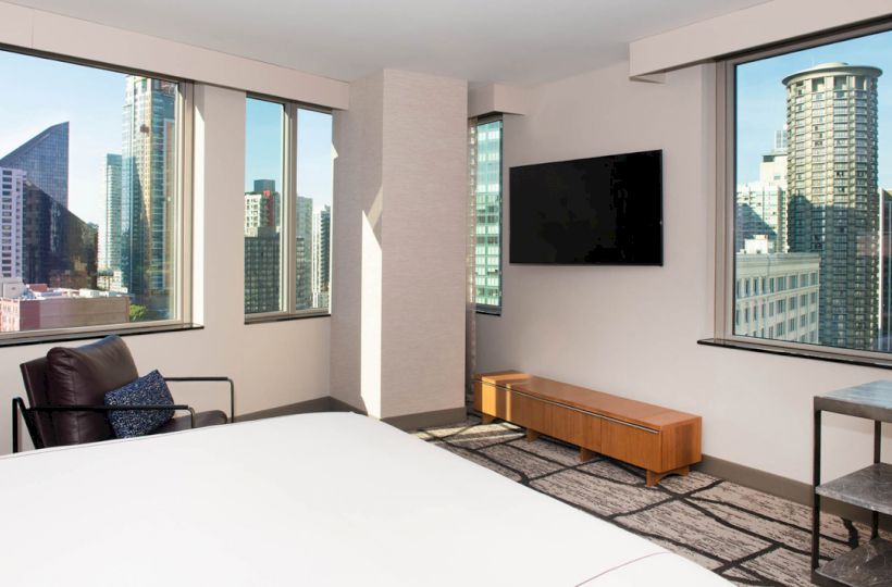 A modern hotel room with a bed, armchair, wall-mounted TV, and large windows showcasing an urban cityscape and skyscrapers.