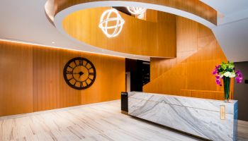 The image shows a modern, elegant reception area with a marble desk, a large wall clock, and contemporary lighting fixtures, with flowers on the desk.