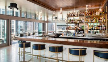 This image shows a stylish bar area with a wooden counter, gold and navy bar stools, shelves stocked with various bottles, and large windows.