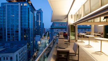 A modern apartment balcony with seating overlooks a city street lined with tall buildings during the evening, illuminated by city lights.