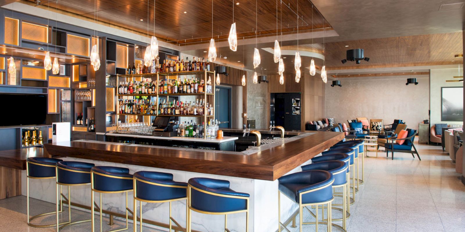 The image shows a modern bar with a central counter, surrounded by blue bar stools, shelves stocked with liquor, and contemporary lighting.