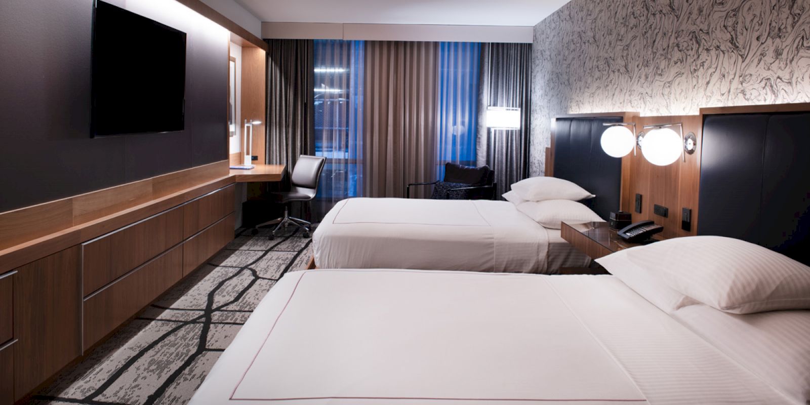 A modern hotel room with two beds, a desk, a chair, a flat-screen TV, and a wall with abstract design. It features subtle lighting and large windows.