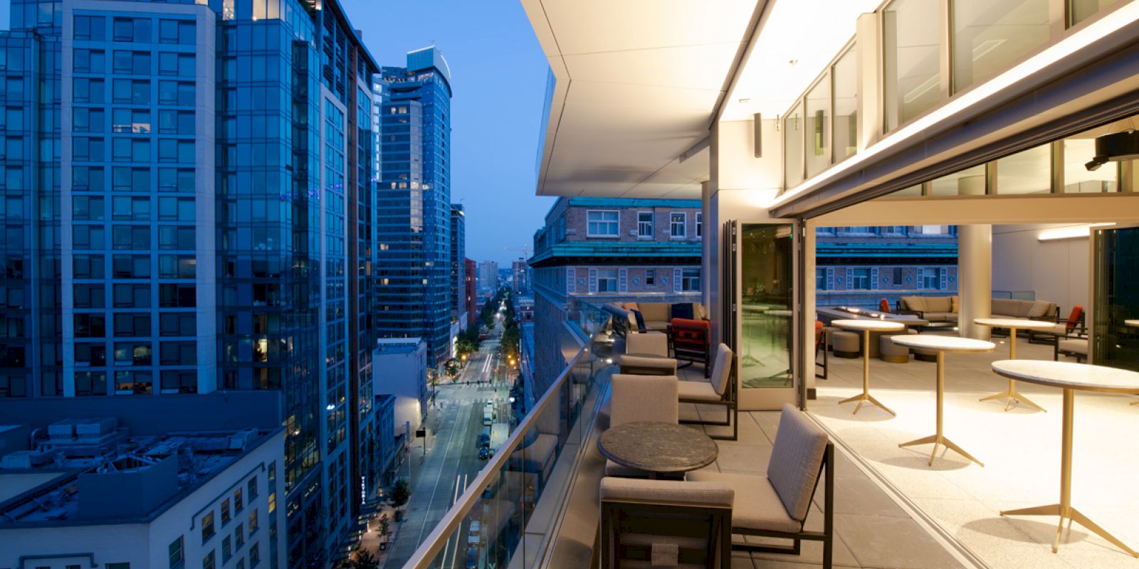 A modern high-rise balcony at dusk, with outdoor seating, city buildings, and a street view illuminated by streetlights and building lights.