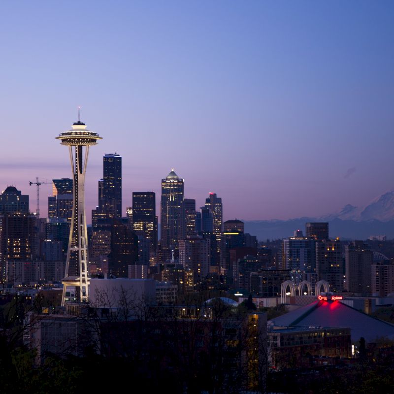 The image shows the Seattle skyline at dusk, featuring the Space Needle prominently with Mount Rainier in the background under a purple and pink sky.
