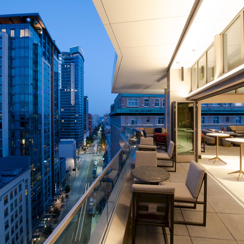The image shows a modern outdoor balcony at dusk with a cityscape view, furnished with tables and chairs, overlooking a busy street below.
