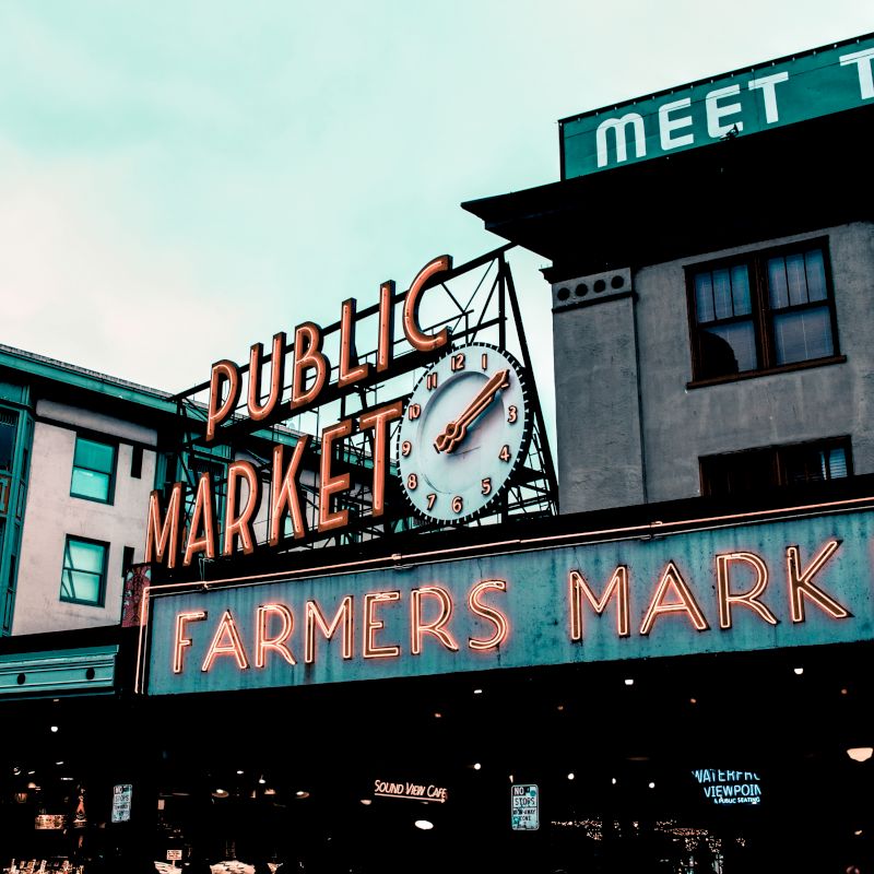 This image shows an iconic neon sign for a Public Market and Farmers Market, with a clock, set against historic buildings on a cloudy day.