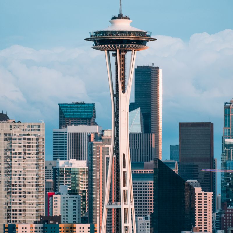 The image shows a cityscape featuring the Space Needle, a famous landmark, with surrounding high-rise buildings under a blue sky with some clouds.