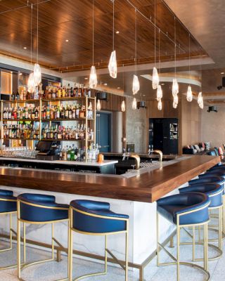 A modern bar with wooden counters, gold-accented chairs, hanging lights, and shelves of liquor bottles, in a spacious setting with tables and couches.