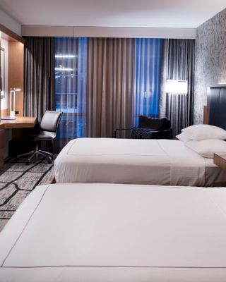 A modern hotel room features two neatly made beds, a wall-mounted TV, a desk with a chair, and stylish decor, under ambient lighting.