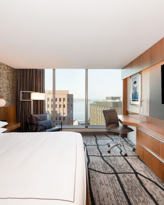 A modern hotel room features a large bed, a desk with a chair, a TV, and floor-to-ceiling windows with a city view, and contemporary decor.