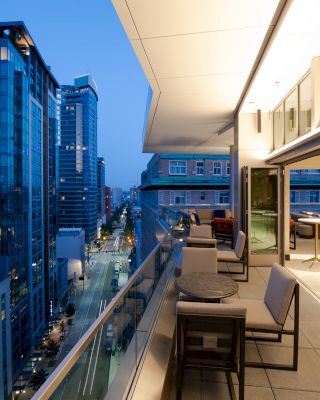 The image shows a modern balcony with tables and chairs, overlooking a cityscape at dusk, featuring tall buildings and a street filled with lights.