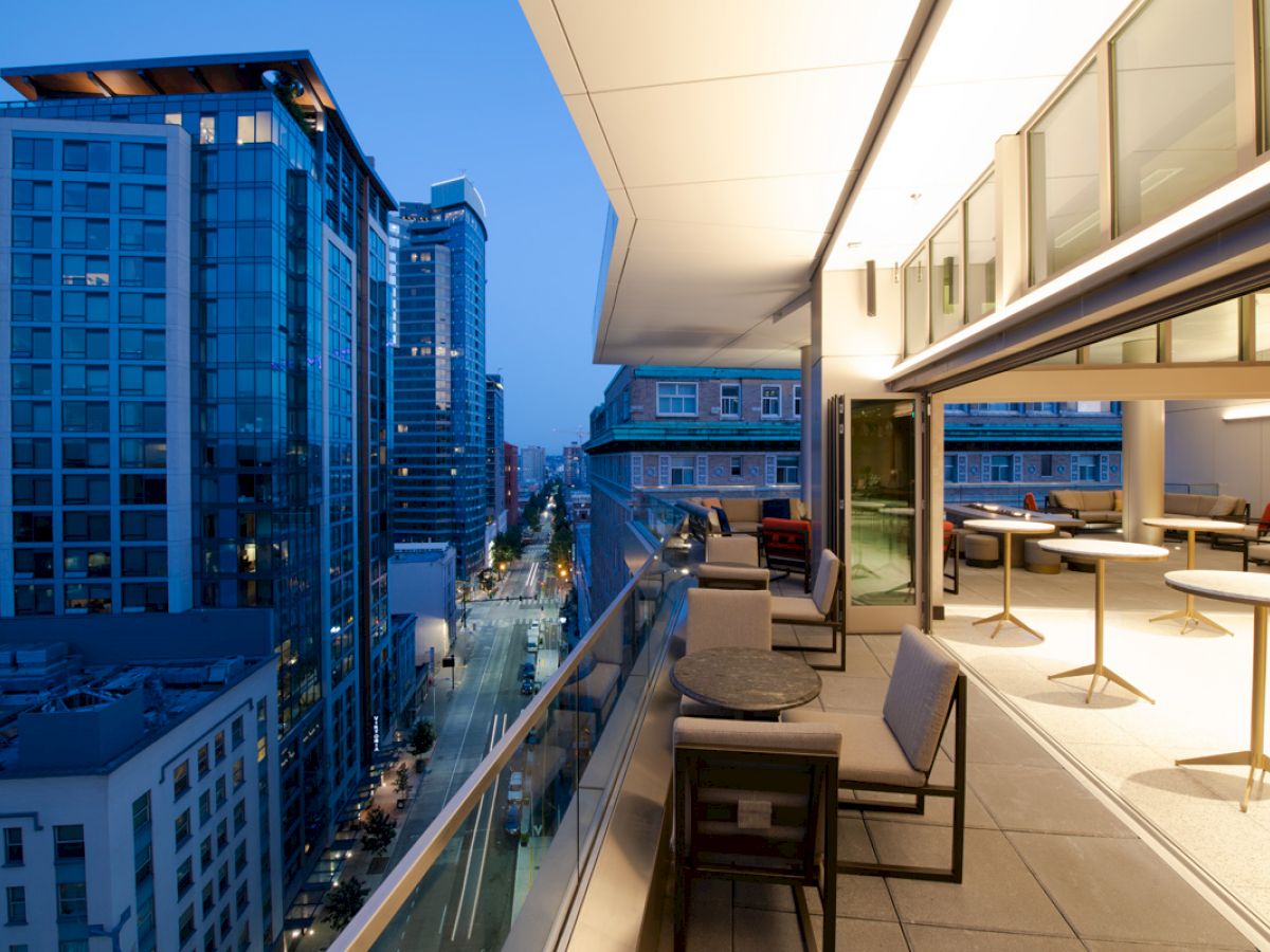 The image shows a cityscape at dusk viewed from a modern balcony with seating and tables, overlooking buildings and a partially lit street.