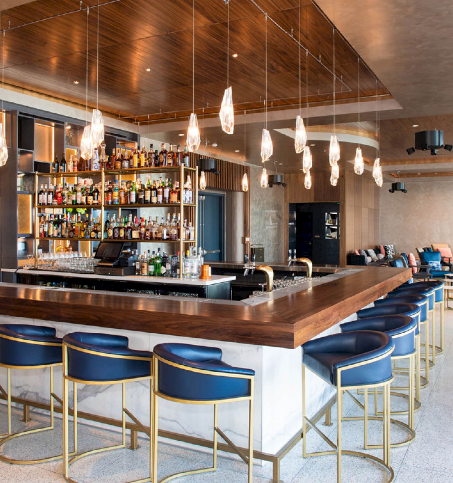 A modern bar with a wooden countertop, blue bar stools, pendant lights, stocked shelves, and seating area in the background.