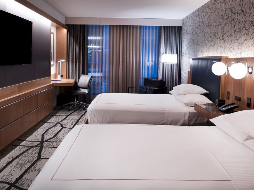 A modern hotel room with two neatly made beds, a desk with a chair, a flat-screen TV on the wall, and a large window with curtains.