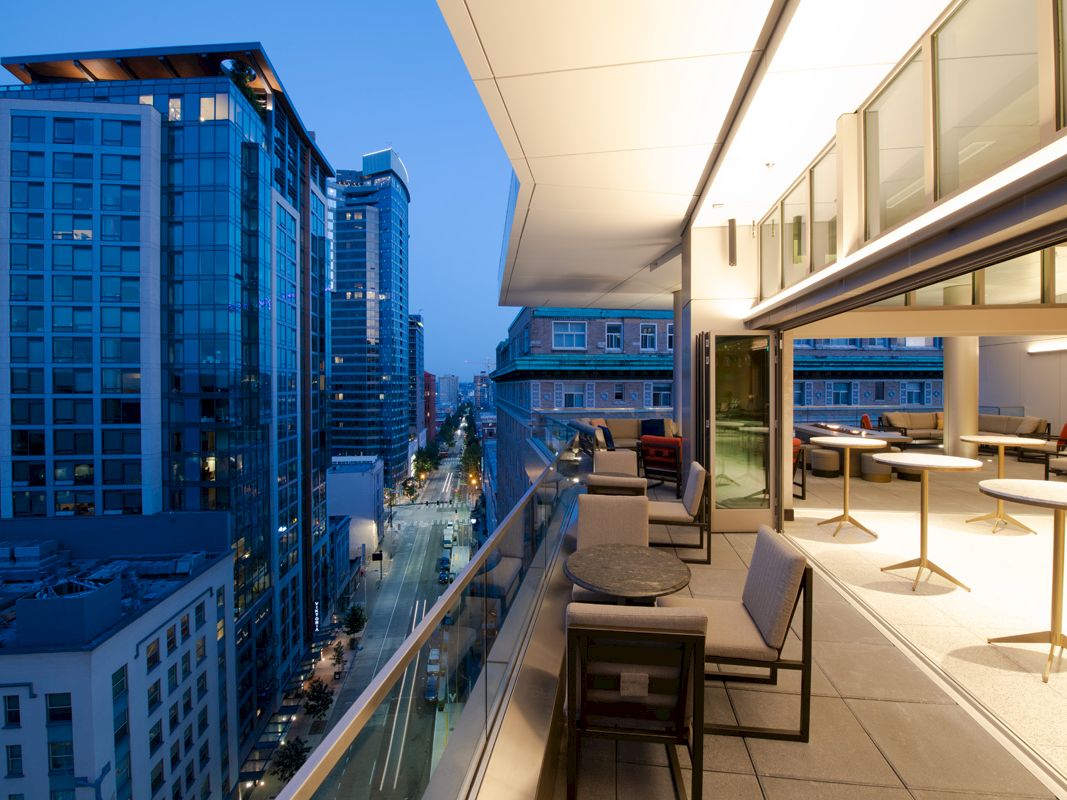 A modern cityscape view from a high-rise balcony with outdoor seating, overlooking lit buildings and a street below during twilight.