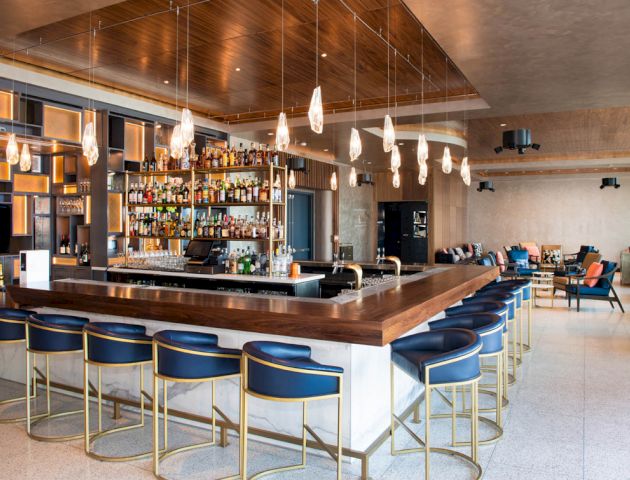 A modern bar with a wooden counter, blue bar stools, hanging lights, and a wide selection of bottles on display behind the counter.