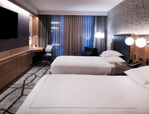 A hotel room with two neatly made beds, a flat-screen TV, a work desk with a chair, and modern lighting and decor.