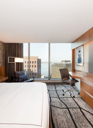 A modern hotel room with a large bed, wall-mounted TV, desk, chair, and floor-to-ceiling windows offering a cityscape view.