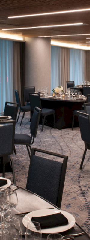 The image shows a banquet hall with round tables set for an event, featuring dark tablecloths, elegant glassware, and floral centerpieces.