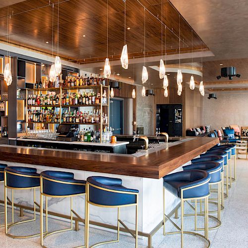 A sleek, modern bar with blue cushioned seats, a well-stocked liquor display, pendant lights, and a spacious seating area in the background.