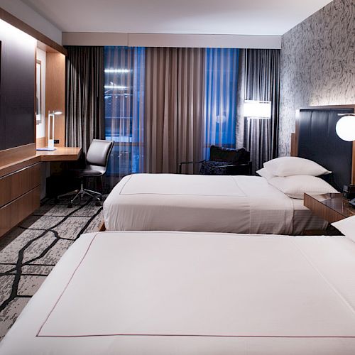 A modern hotel room features two neatly made twin beds, a flat-screen TV on the wall, a work desk with a chair, and contemporary decor.