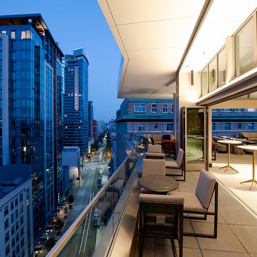 A modern patio with seating and tables overlooks a cityscape of tall buildings, with lights illuminating the scene at dusk or early evening.