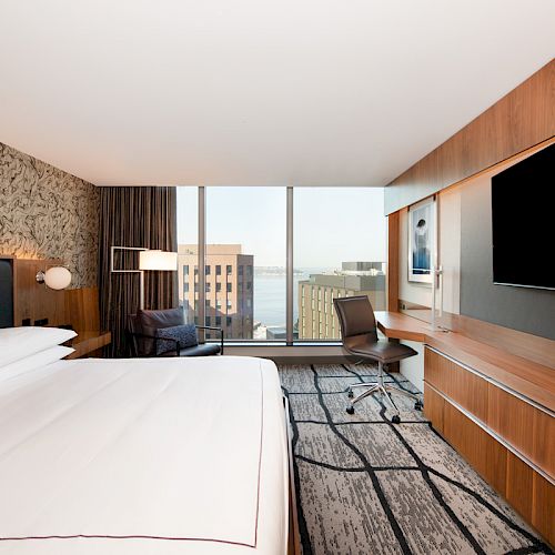 The image shows a modern hotel room with a large bed, desk, TV, and a view of buildings through the window.