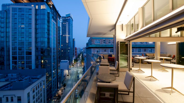 A modern cityscape at dusk featuring high-rise buildings and a furnished balcony with chairs and tables, overlooking a brightly lit street below.