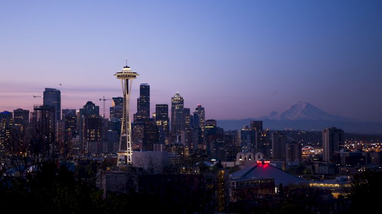 A cityscape of Seattle at dusk featuring the Space Needle, high-rise buildings, and Mount Rainier in the background against a twilight sky.