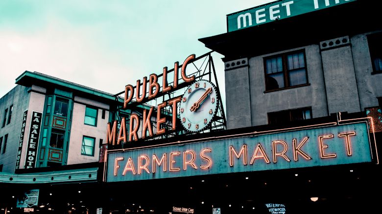 The image shows a public market with a neon sign reading "FARMERS MARKET." A clock is visible above the sign, and buildings surround the area.