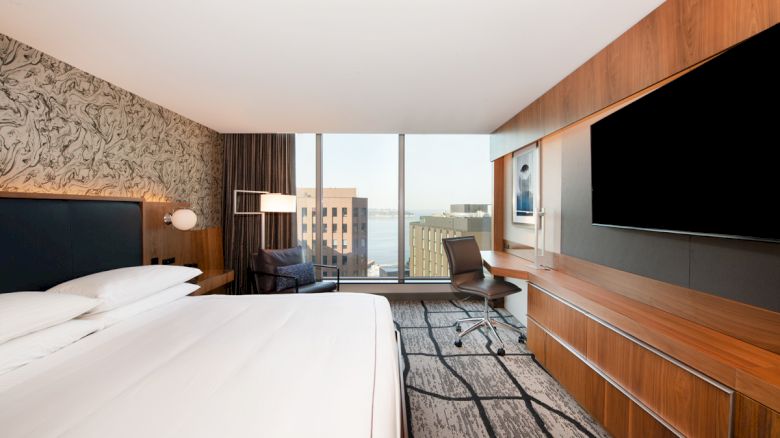 A modern hotel room with a large bed, wall art, a desk and chair, a large TV, and a view of buildings and water through floor-to-ceiling windows.