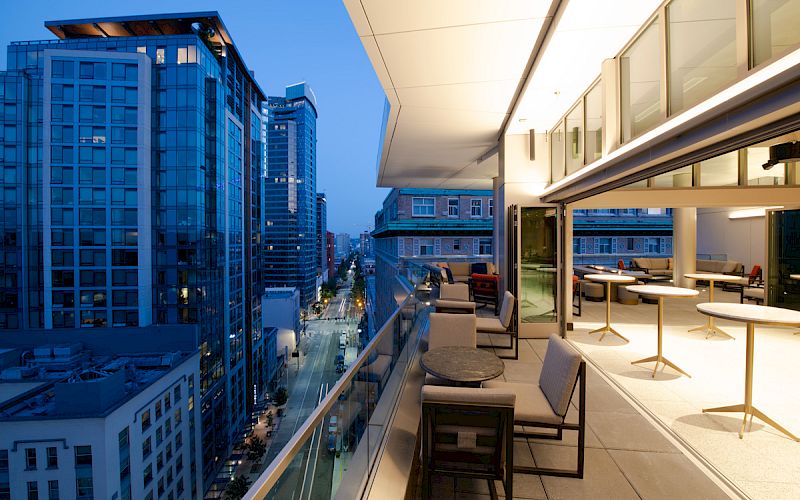 A modern urban balcony at twilight, with seating areas overlooking a cityscape of tall buildings and a street filled with lights.